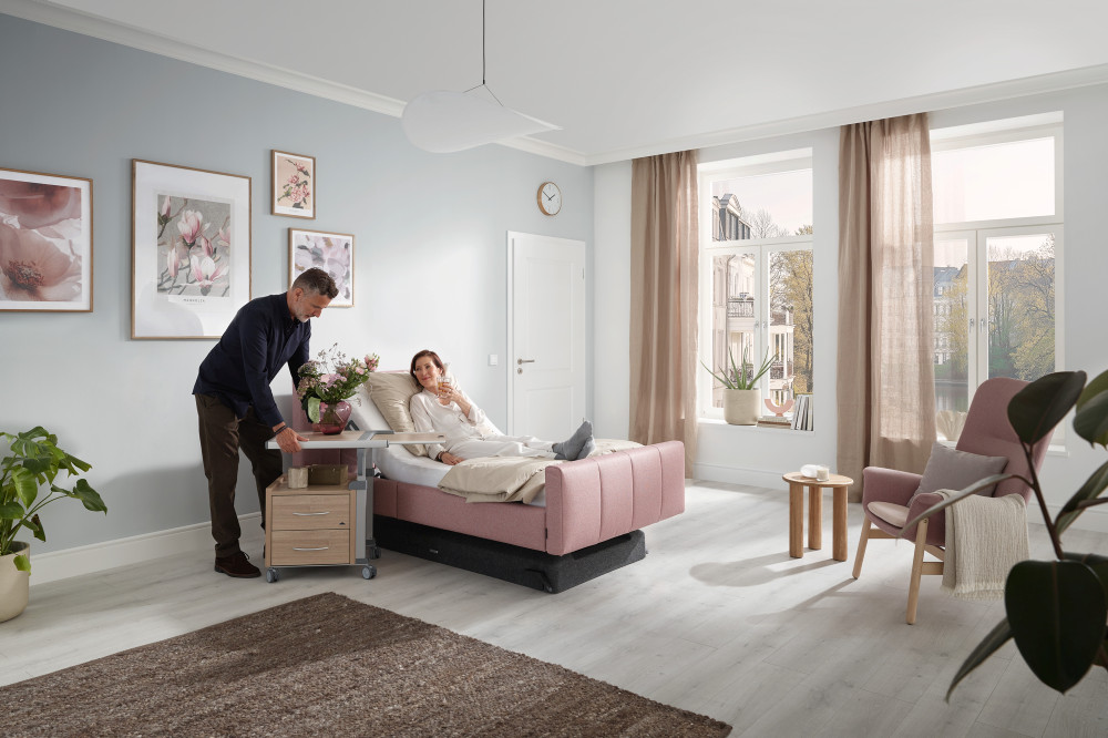 Dame im Komfortbett liegend mit stehendem Mann nebendem Bett